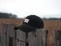 Czarna czapka z daszkiem z napisem "PATRIOTIC" na drewnianym słupku. Czy czapkę z daszkiem można prać w pralce?