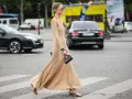 Elegancka, beżowa sukienka maxi i złote sandałki tworzą stylowe beżowa sukienka stylizacje. Kobieta z małą czarną torebką przechodzi przez pasy.