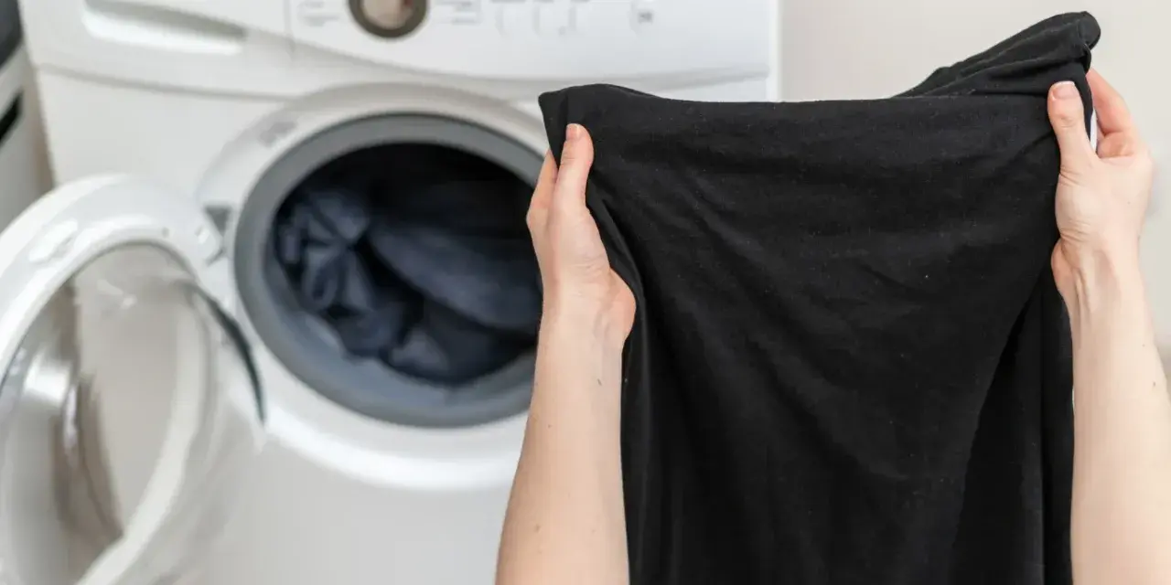 Ręce trzymają czarną koszulkę przed otwartą pralką. Dowiedz się, jak prać czarne ubrania, by zachowały głębię koloru.