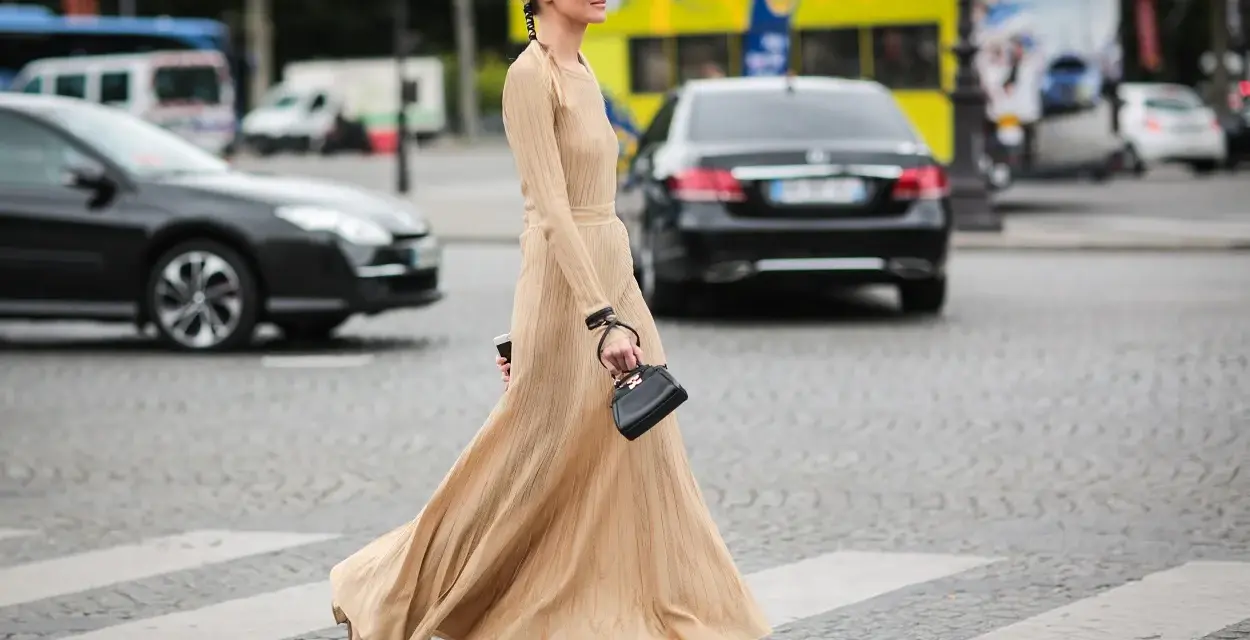 Elegancka, beżowa sukienka maxi i złote sandałki tworzą stylowe beżowa sukienka stylizacje. Kobieta z małą czarną torebką przechodzi przez pasy.