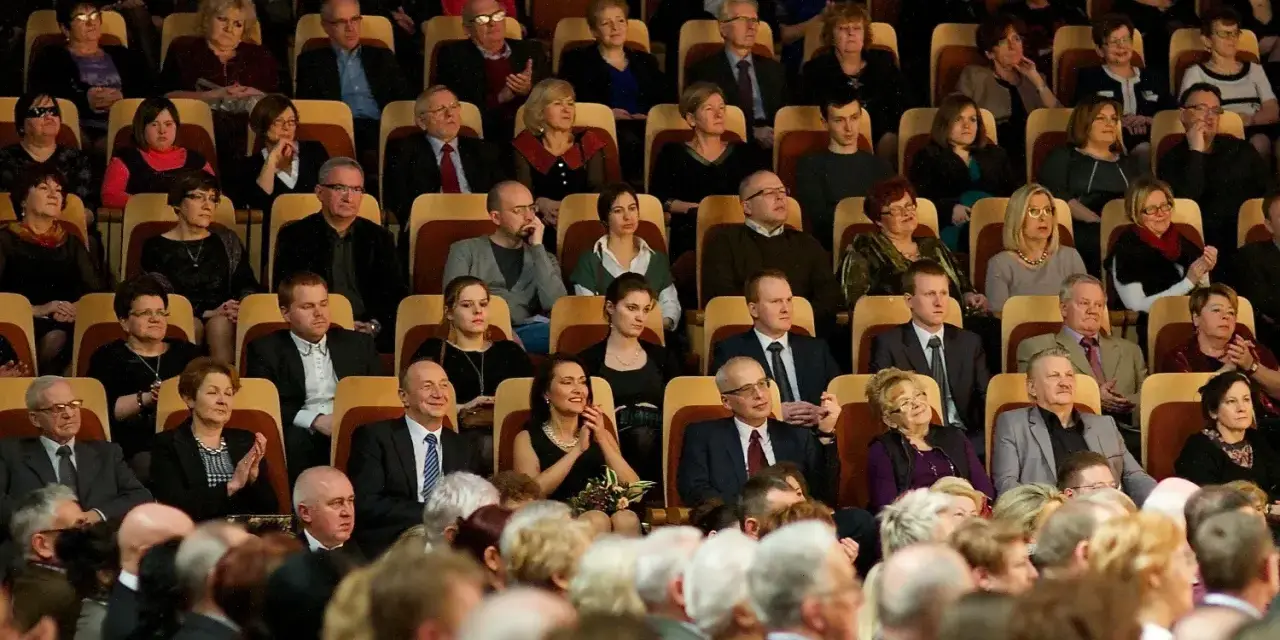 Elegancko ubrani ludzie siedzą w filharmonii, gotowi na koncert. Wskazówka, jak ubrać się do filharmonii: klasyka i szyk.