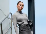 Elegancka kobieta w szarym golfie i jeansach, z czarną torebką, prezentuje styl **business casual** na tle nowoczesnej architektury.