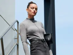 Elegancka kobieta w szarym golfie i jeansach, z czarną torebką, prezentuje styl **business casual** na tle nowoczesnej architektury.