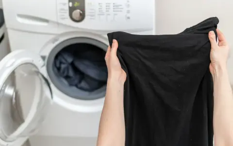 Ręce trzymają czarną koszulkę przed otwartą pralką. Dowiedz się, jak prać czarne ubrania, by zachowały głębię koloru.