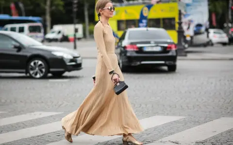 Elegancka, beżowa sukienka maxi i złote sandałki tworzą stylowe beżowa sukienka stylizacje. Kobieta z małą czarną torebką przechodzi przez pasy.