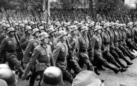 Żołnierze SS maszerują w zwartym szyku, niosąc karabiny. Ich charakterystyczne mundury, których projektant pozostaje nieznany, budzą grozę.