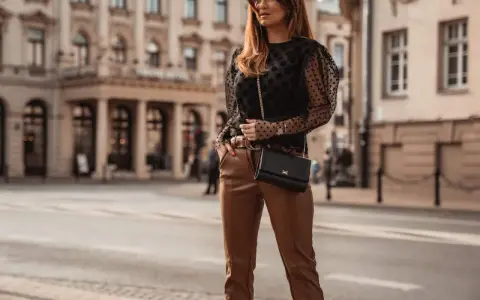 Elegancka kobieta w czarnych, skórzanych spodniach i brązowych butach, z czarną torebką na łańcuszku, pozuje na tle miejskiej architektury.