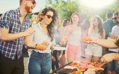 Ludzie cieszą się grillem w słoneczny dzień. Wszyscy ubrani są swobodnie, idealnie na grilla: koszule w kratę, jeansy, szorty.