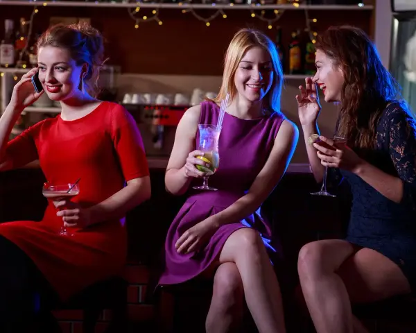 Trzy kobiety w strojach koktajlowych rozmawiają przy barze, pijąc drinki.