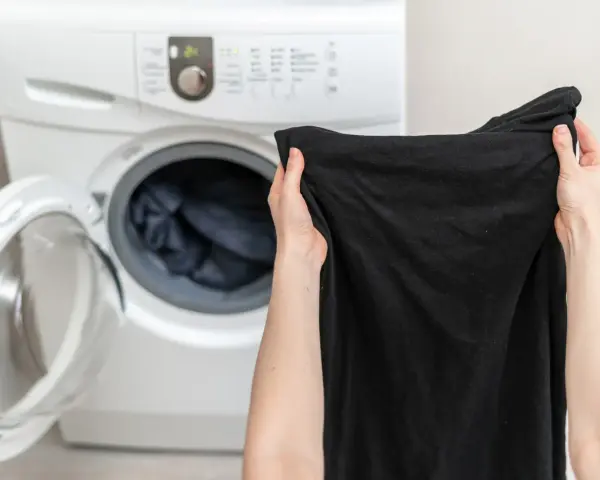 Ręce trzymają czarną koszulkę przed otwartą pralką. Dowiedz się, jak prać czarne ubrania, by zachowały głębię koloru.