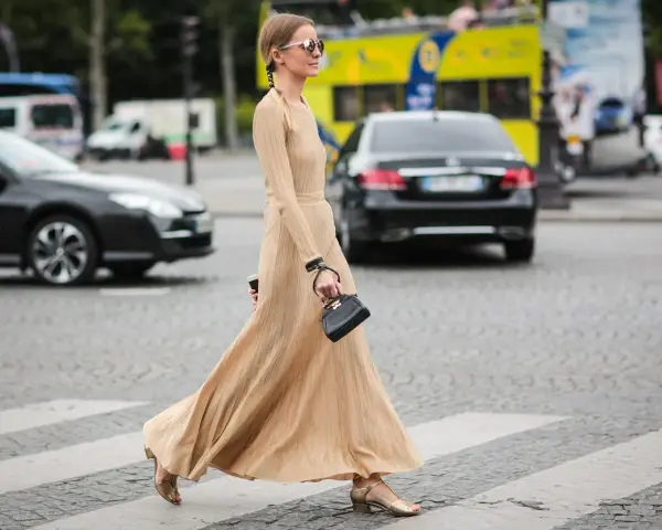 Elegancka, beżowa sukienka maxi i złote sandałki tworzą stylowe beżowa sukienka stylizacje. Kobieta z małą czarną torebką przechodzi przez pasy.
