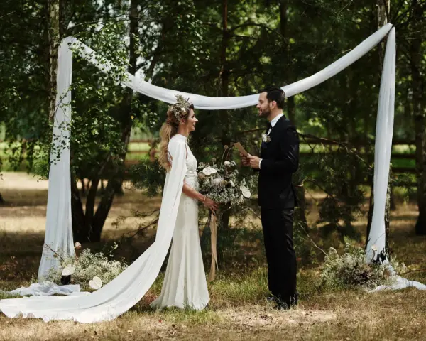 Panna młoda w sukni z peleryną i wiankiem, pan młody w garniturze. Inspiracja, jak się ubrać na wesele w stylu boho.