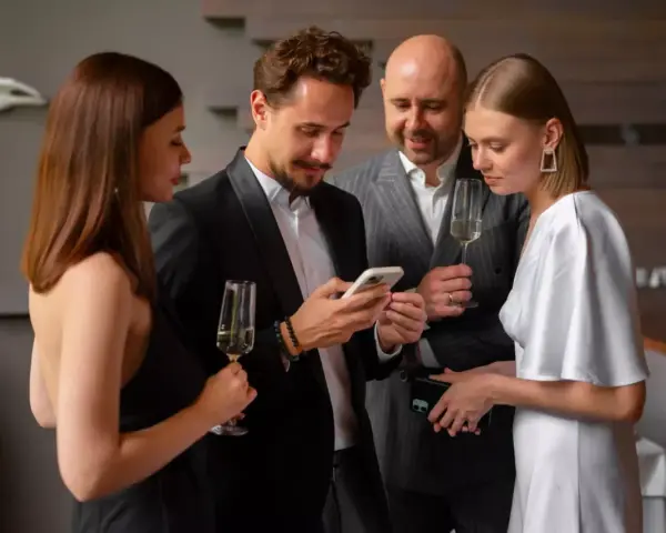 Elegancka kobieta w białej sukni i mężczyzna w czarnym smokingu dyskutują, co założyć na sukienkę wieczorową, trzymając telefony i kieliszki.