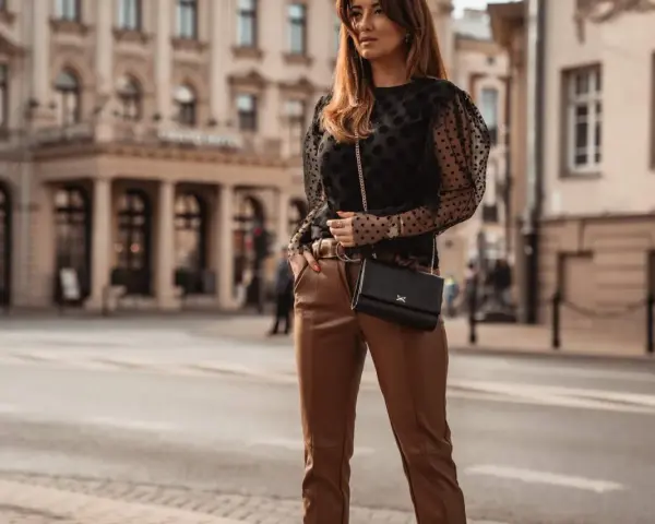 Elegancka kobieta w czarnych, skórzanych spodniach i brązowych butach, z czarną torebką na łańcuszku, pozuje na tle miejskiej architektury.