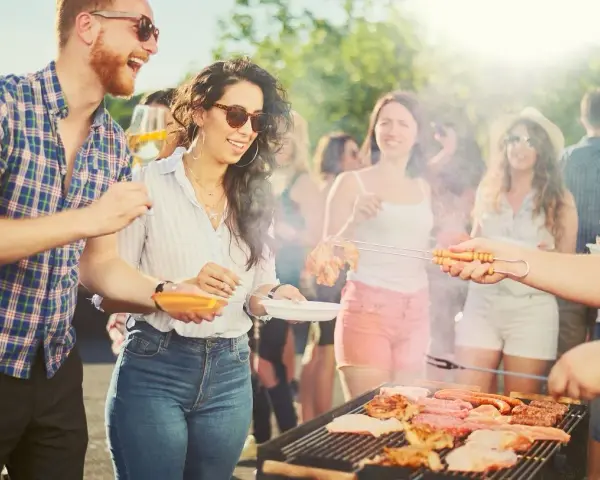 Ludzie cieszą się grillem w słoneczny dzień. Wszyscy ubrani są swobodnie, idealnie na grilla: koszule w kratę, jeansy, szorty.