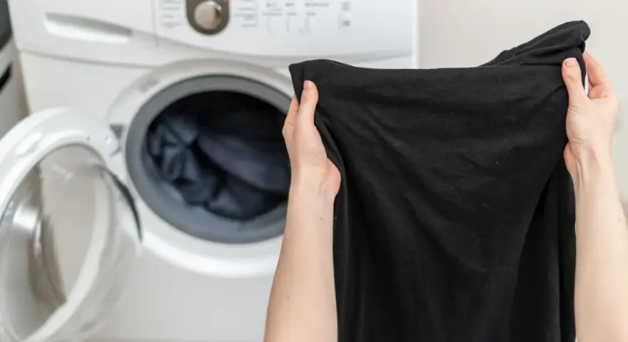 Ręce trzymają czarną koszulkę przed otwartą pralką. Dowiedz się, jak prać czarne ubrania, by zachowały głębię koloru.