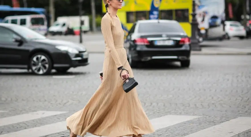 Elegancka, beżowa sukienka maxi i złote sandałki tworzą stylowe beżowa sukienka stylizacje. Kobieta z małą czarną torebką przechodzi przez pasy.