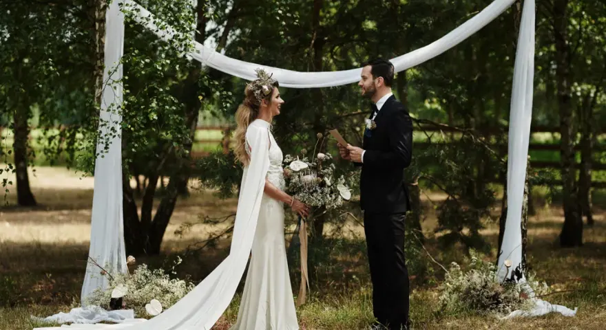 Panna młoda w sukni z peleryną i wiankiem, pan młody w garniturze. Inspiracja, jak się ubrać na wesele w stylu boho.