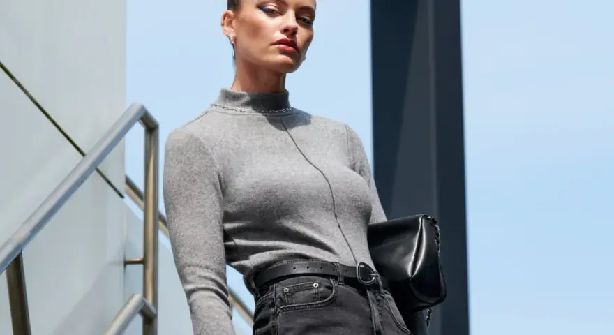 Elegancka kobieta w szarym golfie i jeansach, z czarną torebką, prezentuje styl **business casual** na tle nowoczesnej architektury.