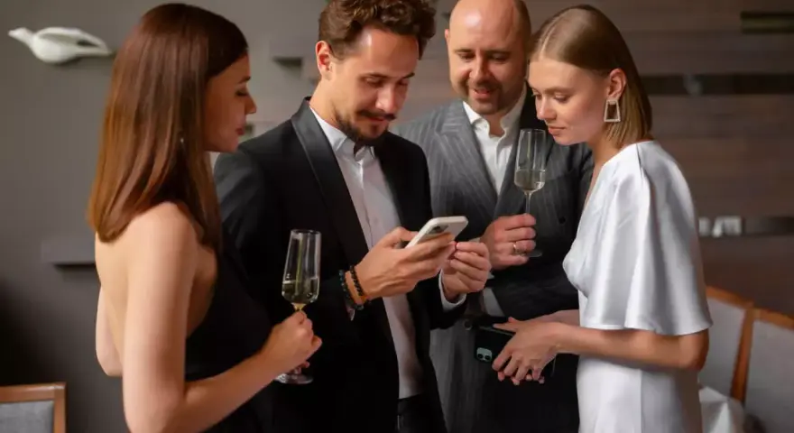 Elegancka kobieta w białej sukni i mężczyzna w czarnym smokingu dyskutują, co założyć na sukienkę wieczorową, trzymając telefony i kieliszki.