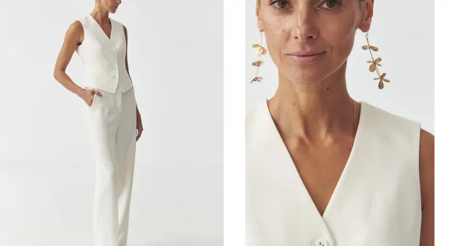 Elegancka kobieta w białej kamizelce bez marynarki i spodniach. Długie, złote kolczyki dodają stylizacji szyku.