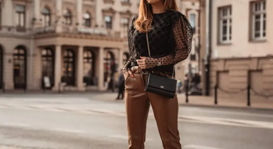 Elegancka kobieta w czarnych, skórzanych spodniach i brązowych butach, z czarną torebką na łańcuszku, pozuje na tle miejskiej architektury.