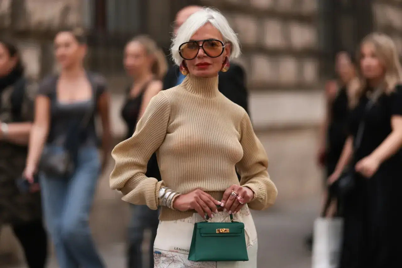 Elegancka kobieta po 60. prezentuje stylowe stylizacje, łącząc beżowy golf z wzorzystą spódnicą i zieloną torebką.